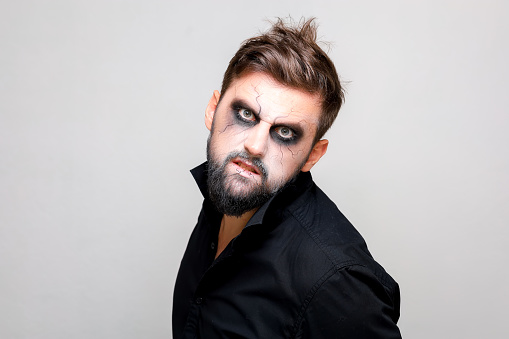 a bearded man with undead-style makeup on Halloween looks with big eyes at the camera