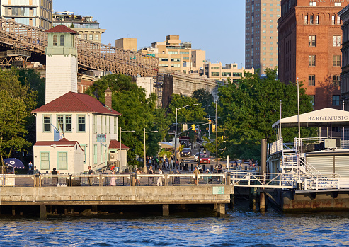 Brooklyn, NY - August 24, 2021: Brooklyn Bridge Park at Fulton Ferry Landing in Brooklyn, NYC, in the early evening on a sunny summer day.The frame building on the left with a red roof was a fireboat station.