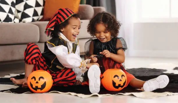 Photo of Two cute african kids brother and sister in Halloween costumes playing together on floor at home