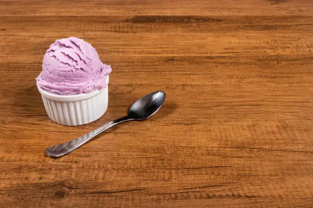Photo of Grape-flavored purple ice cream served in the pot. A ball of ice cream accompanied by a spoon. Left-aligned elements leaving free space for text on the right.