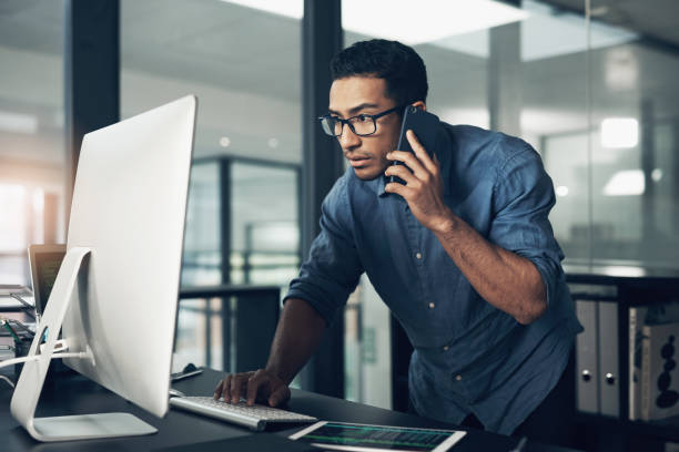 istock Shot of a young man using his cellphone while working on a computer in a modern office 1344688160