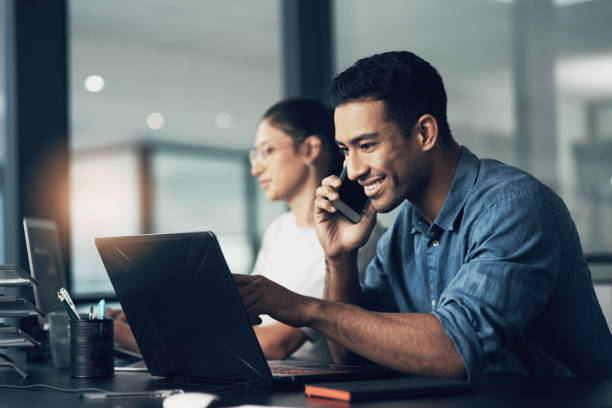 Shot of a young man using his cellphone while working on a laptop in a modern office