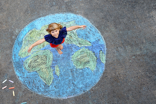 Little preschool girl with earth globe painting with colorful chalks on ground. Positive toddler child. Happy earth day concept. Creation of children for saving world, environment and ecology