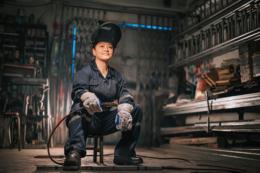 asian chinese female blue collar worker welder with protective workwear looking away smiling in workshop garage sitting on stool