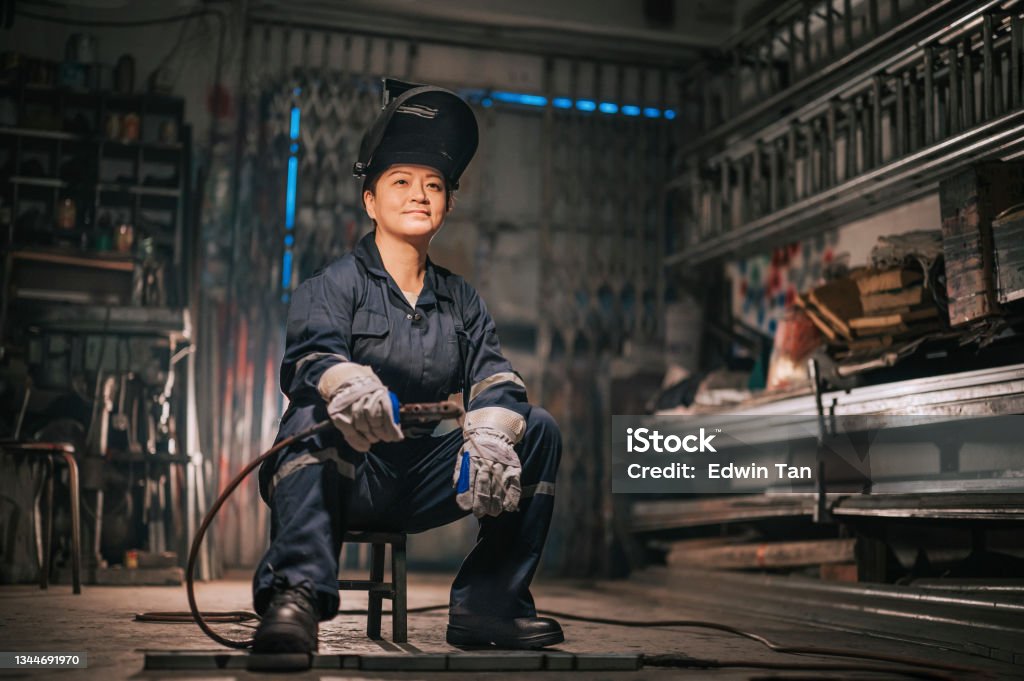 asian chinese female blue collar worker welder with protective workwear looking away smiling in workshop garage sitting on stool Welder Stock Photo