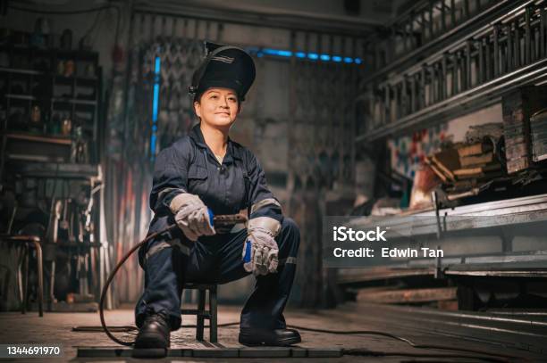 Asian Chinese Female Blue Collar Worker Welder With Protective Workwear Looking Away Smiling In Workshop Garage Sitting On Stool Stock Photo - Download Image Now