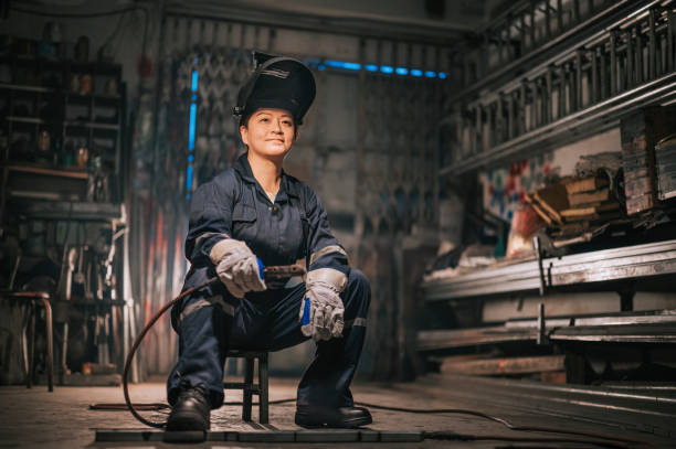 asiática chinesa mulher trabalhador de colarinho azul soldador com roupa de trabalho protetora olhando para longe sorrindo na oficina garagem sentado no banquinho - working class - fotografias e filmes do acervo