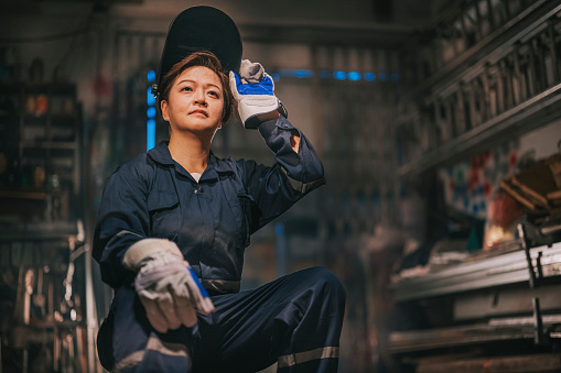 asian chinese female blue collar worker welder with protective workwear looking away smiling in workshop garage sitting on stool