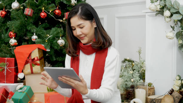 istock Asian woman using smart tablet sit at christmas tree xmas present gift box in cozy living room. Woman hand touch screen smile shopping online, happiness time christmas holiday winter season festive 1344698149