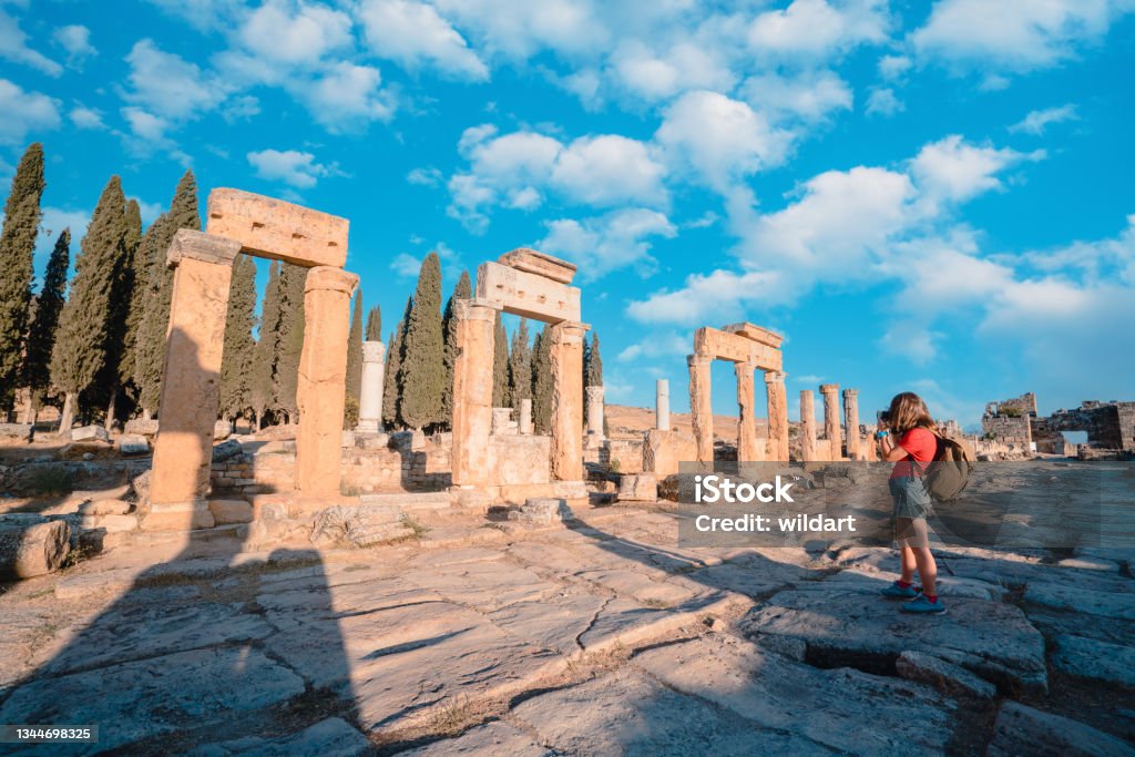 Photographer tourist girl is taking photos of the Frontinus Gate in ancient ruins of Hierapolis , Pamukkale UNESCO, Backpacker, Camera, Travertine pools, Greek architecture Türkiye - Country Stock Photo