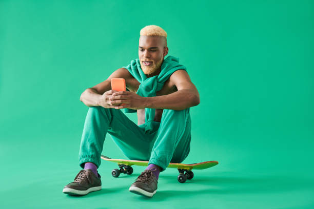 Man with naked torso using mobile phone and smiling while sitting Chatting with you. Full length view of young man with naked torso using mobile phone and smiling while sitting at his skateboard. Isolated on green background black guy with blonde hair stock pictures, royalty-free photos & images
