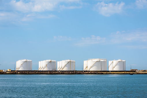 Large white storage tanks for oil and fuel in a marine oil terminal under a blue sky.