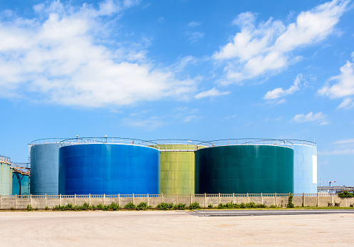 Large coloured storage tanks for oil and fuel in a tank farm under a blue sky with white clouds.