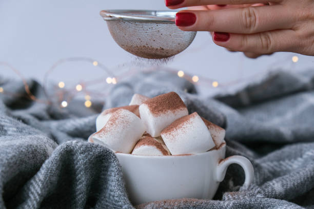 istock Adding cacao powder on top of hot chocolate with marshmallows in white cup on sweater background. Cozy christmas lights 1344704054