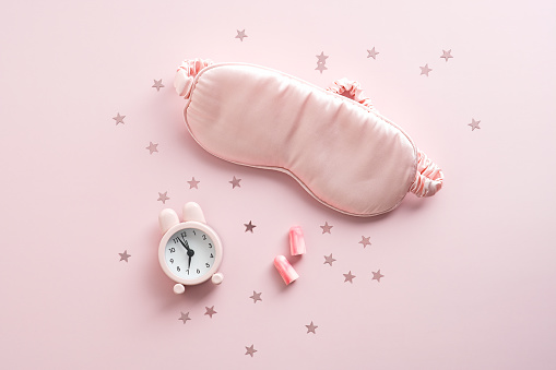 Sleeping mask, alarm clock, earplugs and glitter on pink background. Bedtime accessory. Flat lay, top view.