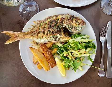 Horizontal close up plate of local caught fresh while snapper fish as a seafood lunch with thick cut chips pear amd pine nut rocket salad dining al fresco next to ocean waves at jetty promenade Coffs Harbour Australia
