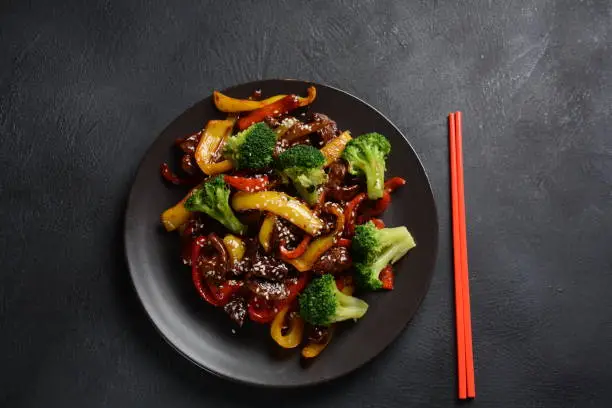Asian teriyaki beef with red and yellow bell peppers, broccoli and sesame seeds on a plate on the table. Spicy teriyaki beef stir fry with vegetables and rice  on a dark background