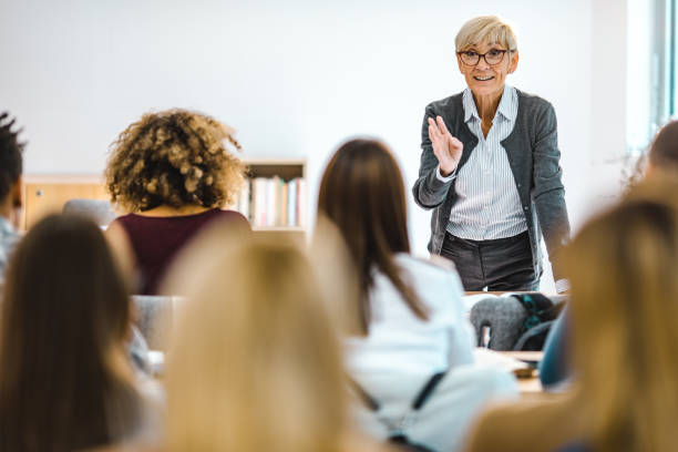 Happy mature professor giving a lecture in front of her students at lecture hall. Happy senior teacher talking to large group of college students in amphitheater. professor stock pictures, royalty-free photos & images