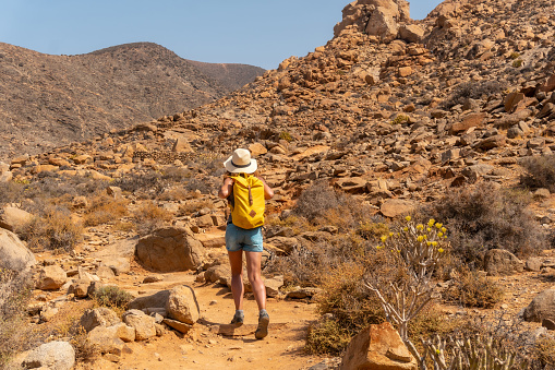 A hiker with a hat and yellow backpack walking along the canyon path towards the Mirador de la Peñitas, Fuerteventura, Canary Islands. Spain