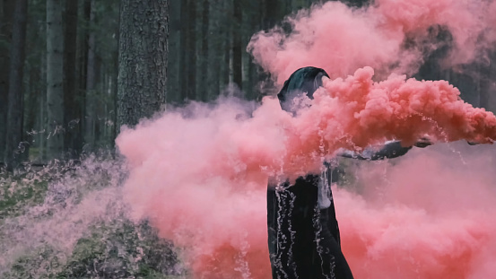 A mysterious man in a raincoat with a smoke bomb in his hand. The witch smokes in the forest with magical pink smoke. Mystical background.