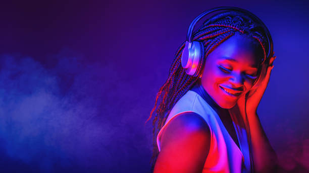 Stylish fashion african american teenager model wearing headphones listening dj music dancing in purple neon lights. Young teen girl enjoy cool music stock photo