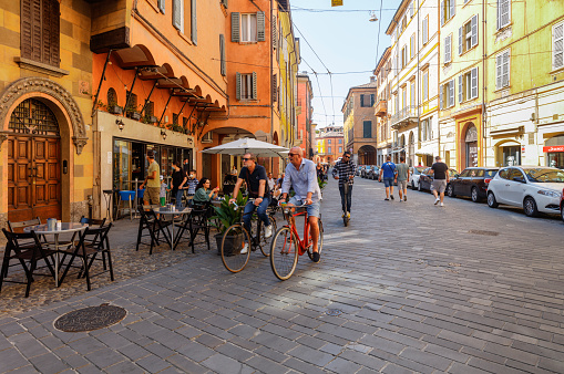 Modena, Italy - September 4, 2021: Street in the old town, with people on bicycles and customers sitting at the tables of an outdoor restaurant.