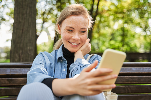 Portrait view of the happy woman sitting at the bench and using her smartphone while walking at the park during the warm day. People and technologies concept
