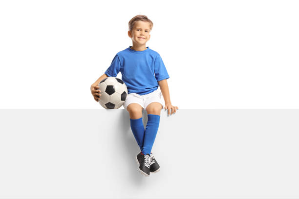 istock Cute little boy in a blue soccer jersey sitting on a blank panel and smiling at camera 1344728334