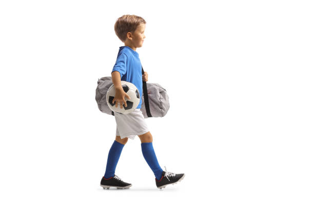 photo de profil pleine longueur d’un garçon portant un maillot de sport, tenant un ballon et un sac de football et marchant - foot walk photos et images de collection