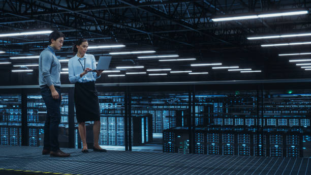istock Data Center Female System Administrator and Male IT Specialist Talk, Use Laptop Computer. Information Technology Engineers work on Cyber Security Network Protection in Cloud Computing Server Farm. 1344731969