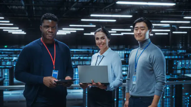 Photo of Diverse Team of Data Center System Administrators and IT Specialists Use Laptop and Tablet Computers, Smiling on Camera. IT Engineers work on Cyber Security Protection in Cloud Computing Server Farm.