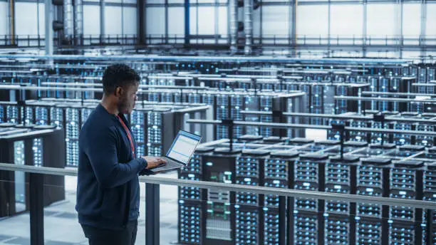 Photo of Data Center Engineer Using Laptop Computer. Server Farm Cloud Computing Specialist Facility with African American Male System Administrator Working with Data Protection Network for Cyber Security.