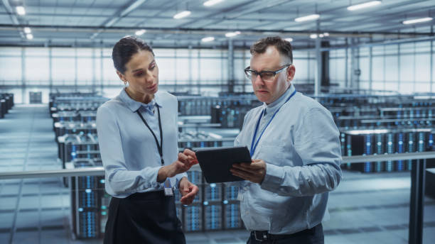 istock Data Center Female System Administrator and Male IT Specialist Talk, Use Tablet Computer. Information Technology Engineers work on Cyber Security Network Protection in Cloud Computing Server Farm. 1344732037