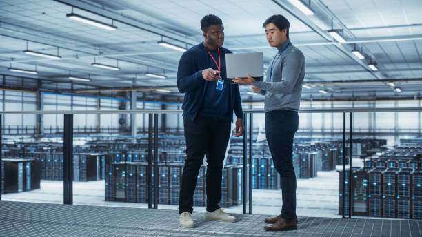 istock Two Multiethnic Male Data Center System Administrators and IT Specialists Talk, Use Laptop. Information Technology Engineers work on Cyber Security Network Protection in Cloud Computing Server Farm. 1344732049