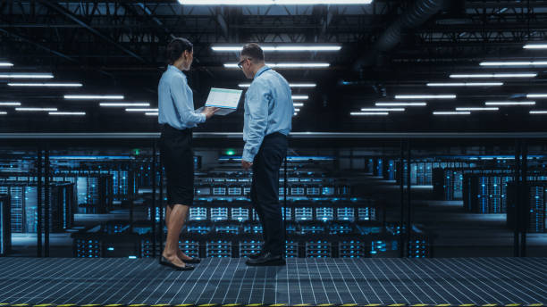 istock Data Center Female System Administrator and Male IT Specialist Talk, Use Laptop Computer. Information Technology Engineers work on Cyber Security Network Protection in Cloud Computing Server Farm. 1344732150