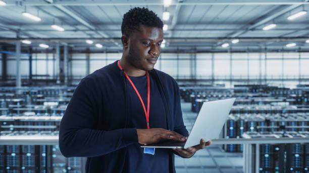 retrato de un ingeniero de centro de datos usando una computadora portátil. instalación especializada en sala de servidores con administrador de sistemas masculino afroamericano que trabaja con la red de protección de datos para la seguridad cibernétic - network server black computer network rack fotografías e imágenes de stock