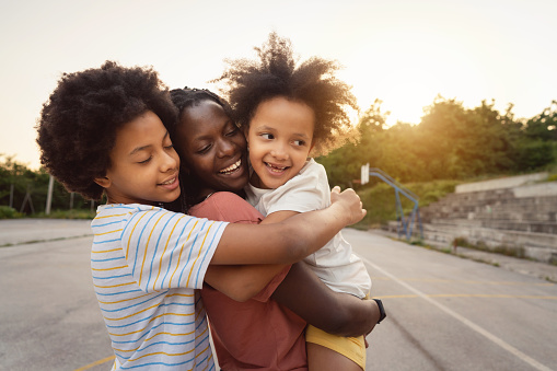 Adorable and cute sisters age 11 and 5, mixed-race ethnicity, embracing their mother of Black ethnicity, bonding and sharing affectionate during their recreational pursuit at the sport court