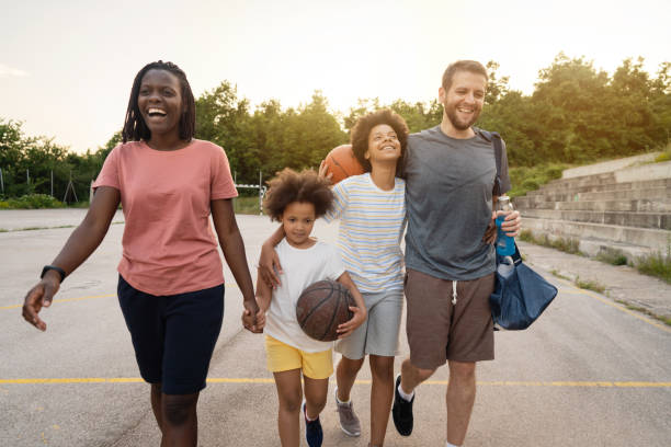 Cheerful blended family, leaving the sports court while holding hands Lovely happy multiracial stepfamily, leaving the sports court after they finished with their family outdoor sports activity, basketball game multiracial person stock pictures, royalty-free photos & images