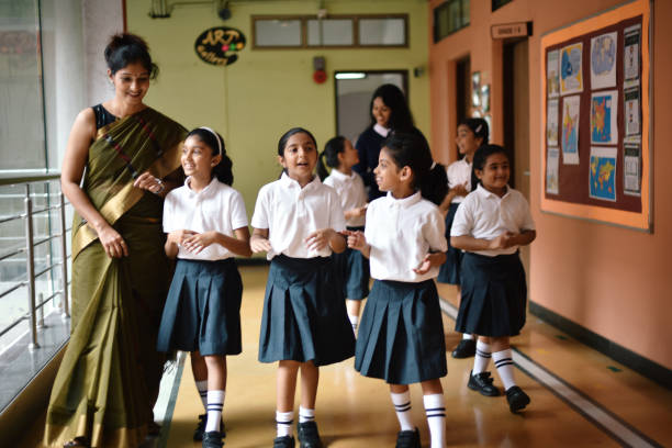Two indian teenage boy school students in uniform sitting at desk in classroom, studying and discussing about their writing assignment by looking at the notebook during class lesson and one other student sitting in the foreground reading a book. Indian students and secondary education concept.