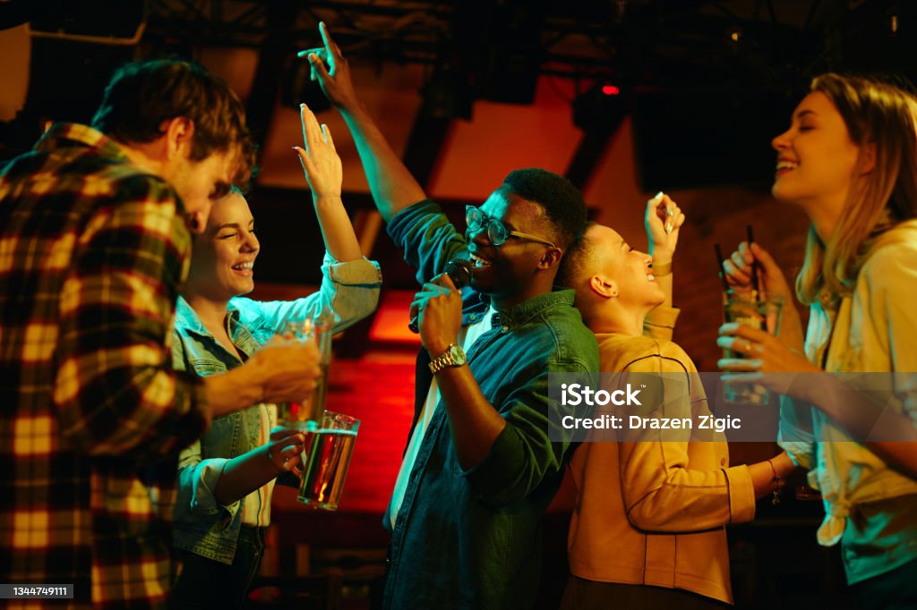 Happy black man singing while being on karaoke party with his friends in a pub. Multi-ethnic group of young people having fun and singing karaoke in a bar at night. Focus is on black man. Karaoke Stock Photo