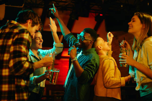 Happy black man singing while being on karaoke party with his friends in a pub. Multi-ethnic group of young people having fun and singing karaoke in a bar at night. Focus is on black man. karaoke stock pictures, royalty-free photos & images