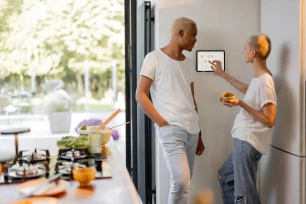 European girl choosing temperature on thermostat of smart home system while her black boyfriend looking at her. Concept of modern technologies in domestic lifestyle. Interior of kitchen.