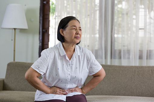 Elderly Asian woman sitting and having a stomachache at home. Retirement, quarantine and health care concept
