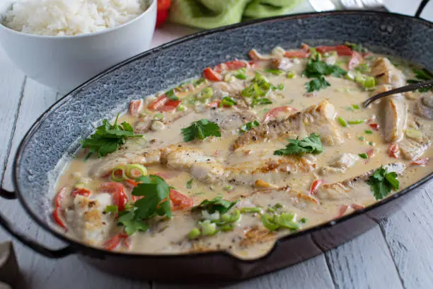 Delicious lactose free and gluten free meal with pan fried redfish fillet cooked with bell peppers, scallions, garlic, lime and ginger in coconut milk. Served with rice on a white wooden table. Closeup from above