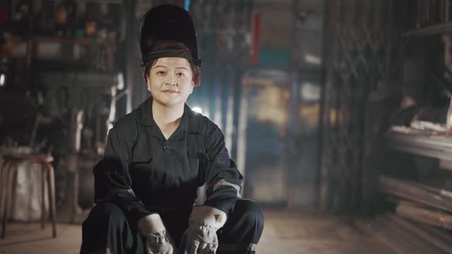 asian chinese female blue collar worker welder with protective workwear looking at camera smiling in workshop garage sitting on stool