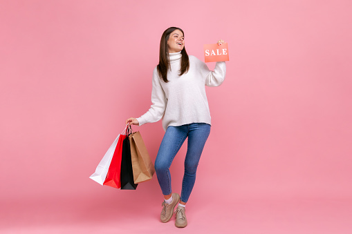 Full length portrait of smiling young adult female holding shopping bags and card with sale inscription, wearing white casual style sweater. Indoor studio shot isolated on pink background.