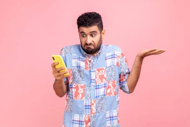 Portrait of attractive bearded man wearing blue casual shirt holding smart phone in hands, spreading hans aside, dont know how to use application. Indoor studio shot isolated on pink background.