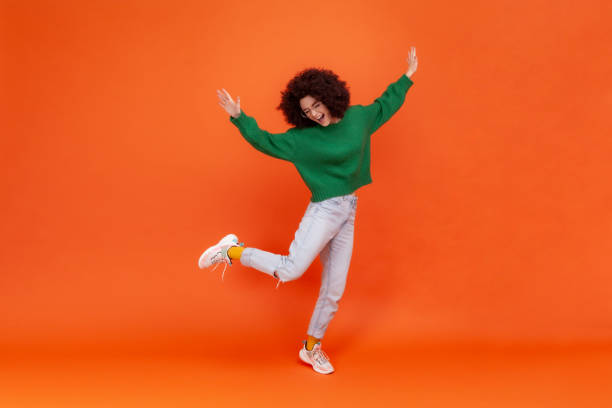 retrato de cuerpo entero de mujer feliz con peinado afro con suéter verde de estilo casual de pie sobre una pierna, brazos levantados, bailando, celebrando. - dacing fotografías e imágenes de stock