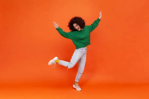 Full length portrait of happy woman with Afro hairstyle wearing green casual style sweater standing on one leg, raised arms, dancing, celebrating. Indoor studio shot isolated on orange background.