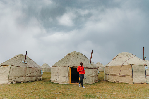 Young male traveler in red shirt and eyeglasses drinking coffee from a cup staying near the traditional yurt at the campsite looking at the scenic lake Song Kul in Kyrgyzstan